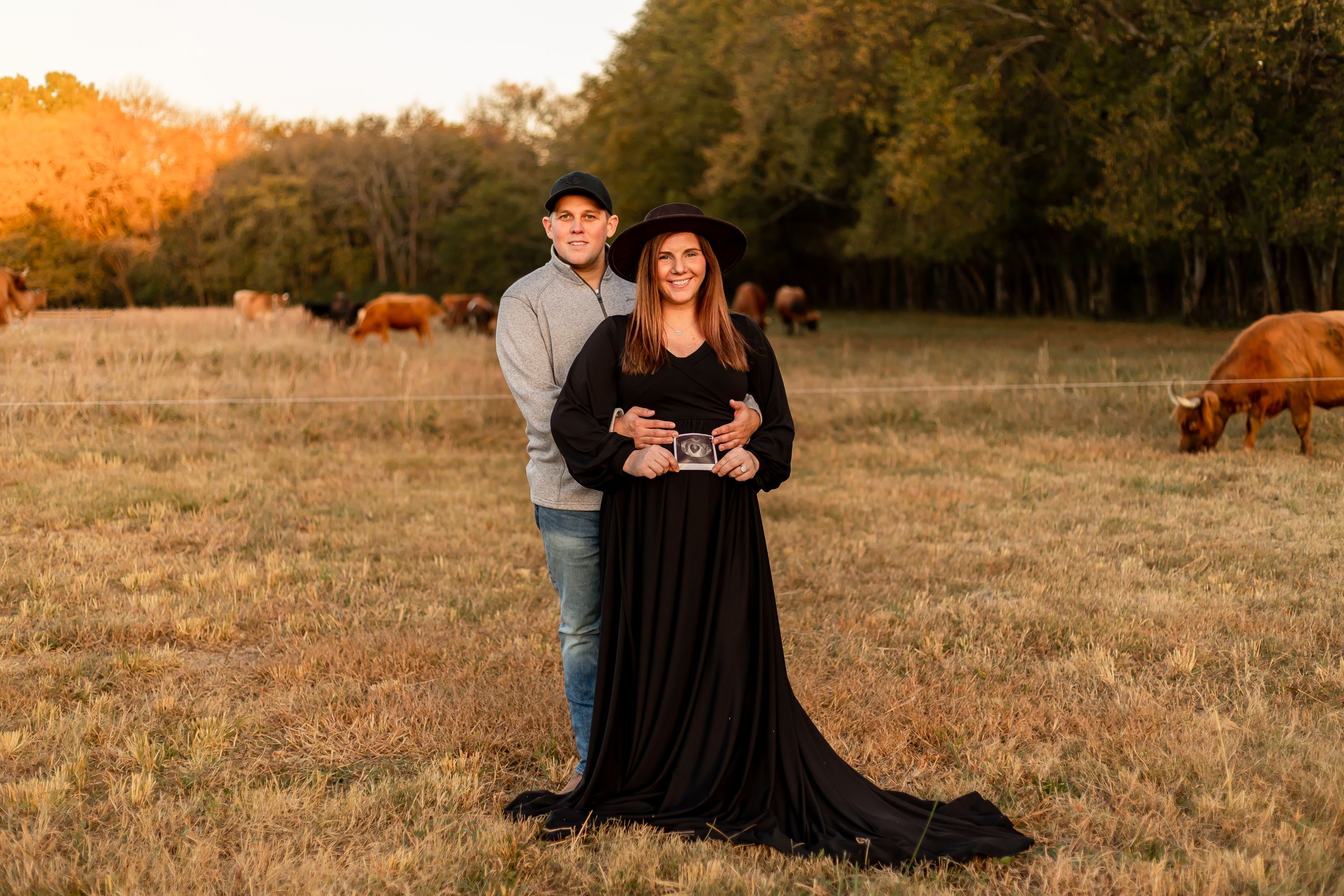 A couple stands together in a field at golden hour with grazing cows in the background.