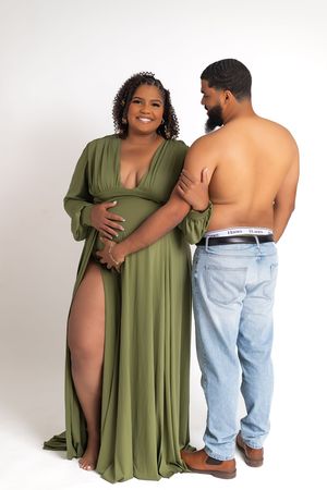 Pregnant woman in olive green maternity gown poses with shirtless partner in jeans for maternity photoshoot.