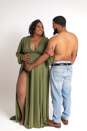 Pregnant woman in olive green maternity gown poses with shirtless partner in jeans during studio maternity photoshoot.