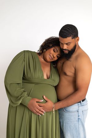 A pregnant woman in a green dress and shirtless man in jeans share a tender maternity photo moment.