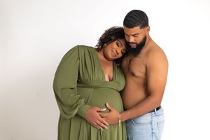 Pregnant woman in green dress with shirtless partner embracing her from behind in a maternity photo shoot.