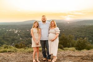 Pregnant woman poses with man and young girl at golden hour overlook with scenic mountain valley view.