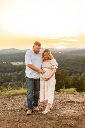 Couple sharing a tender maternity moment at golden hour on a hilltop with scenic mountain views in the background.