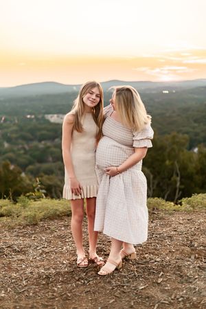 A pregnant woman and a young girl pose together at sunset on a hilltop with scenic mountain views.