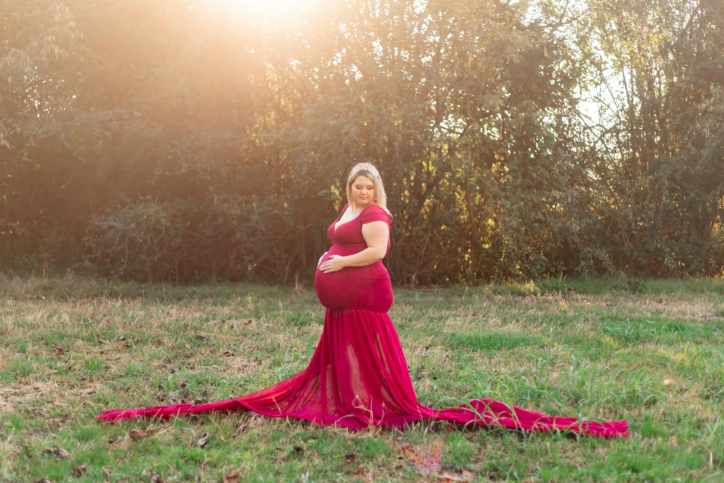 Expecting mother in flowing red gown poses gracefully in sunlit field during golden hour maternity photoshoot.