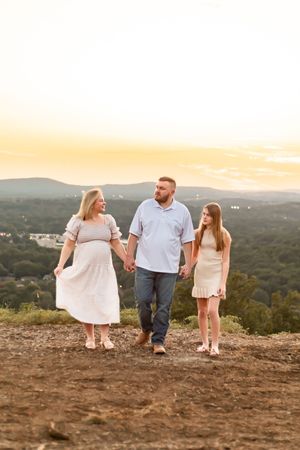 Pregnant woman in white dress walks hand-in-hand with man and girl at golden hour on hilltop with mountain views.