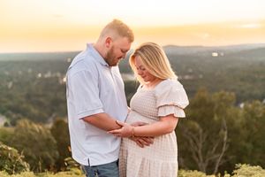 Couple tenderly holding pregnant belly at golden hour sunset with scenic landscape in background.