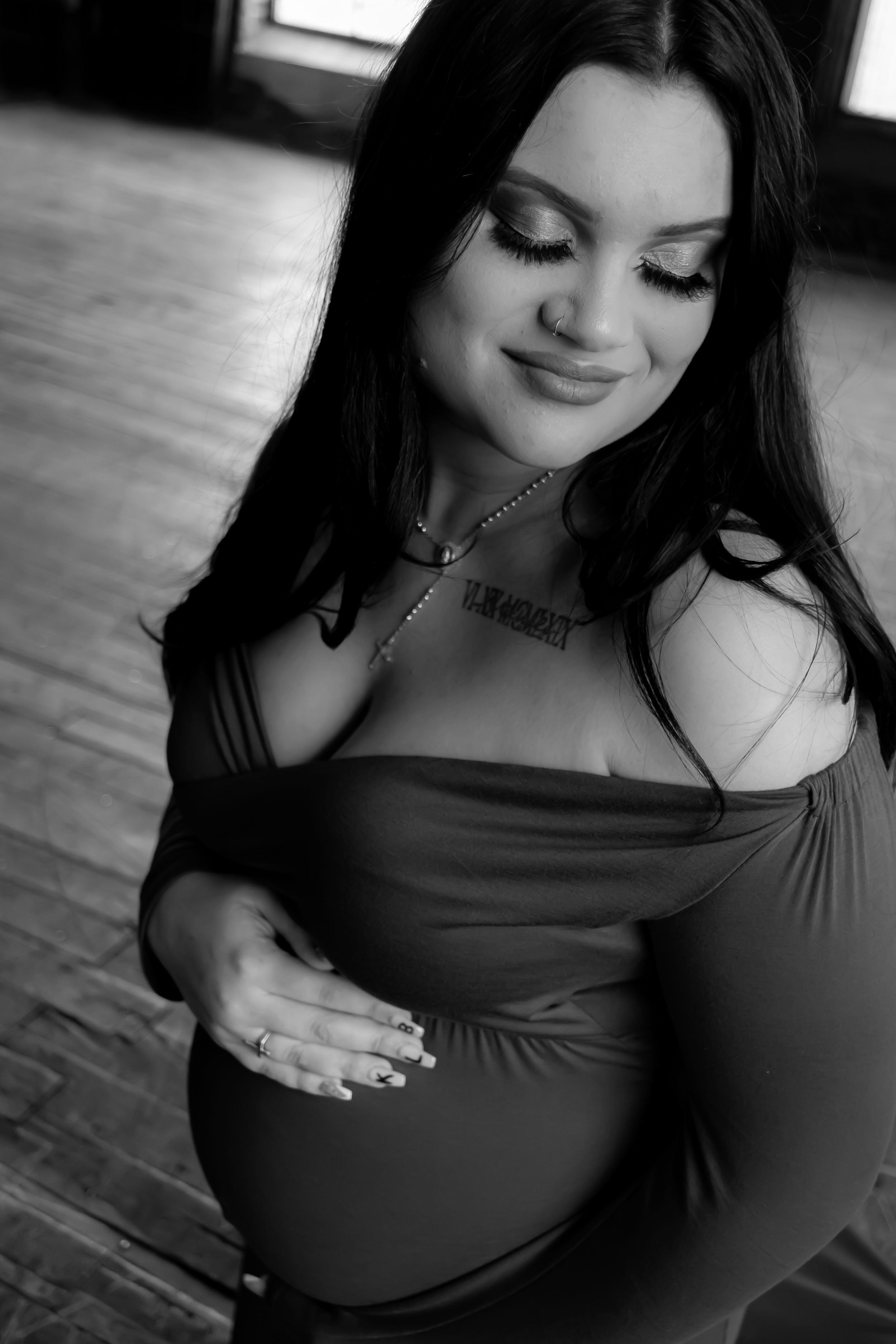 Black and white maternity portrait showing a woman in dark clothing touching her pregnant belly while looking down with a smile.