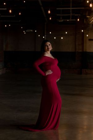A figure in an elegant red long-sleeve maternity gown poses in a dark atmospheric room with hanging string lights.