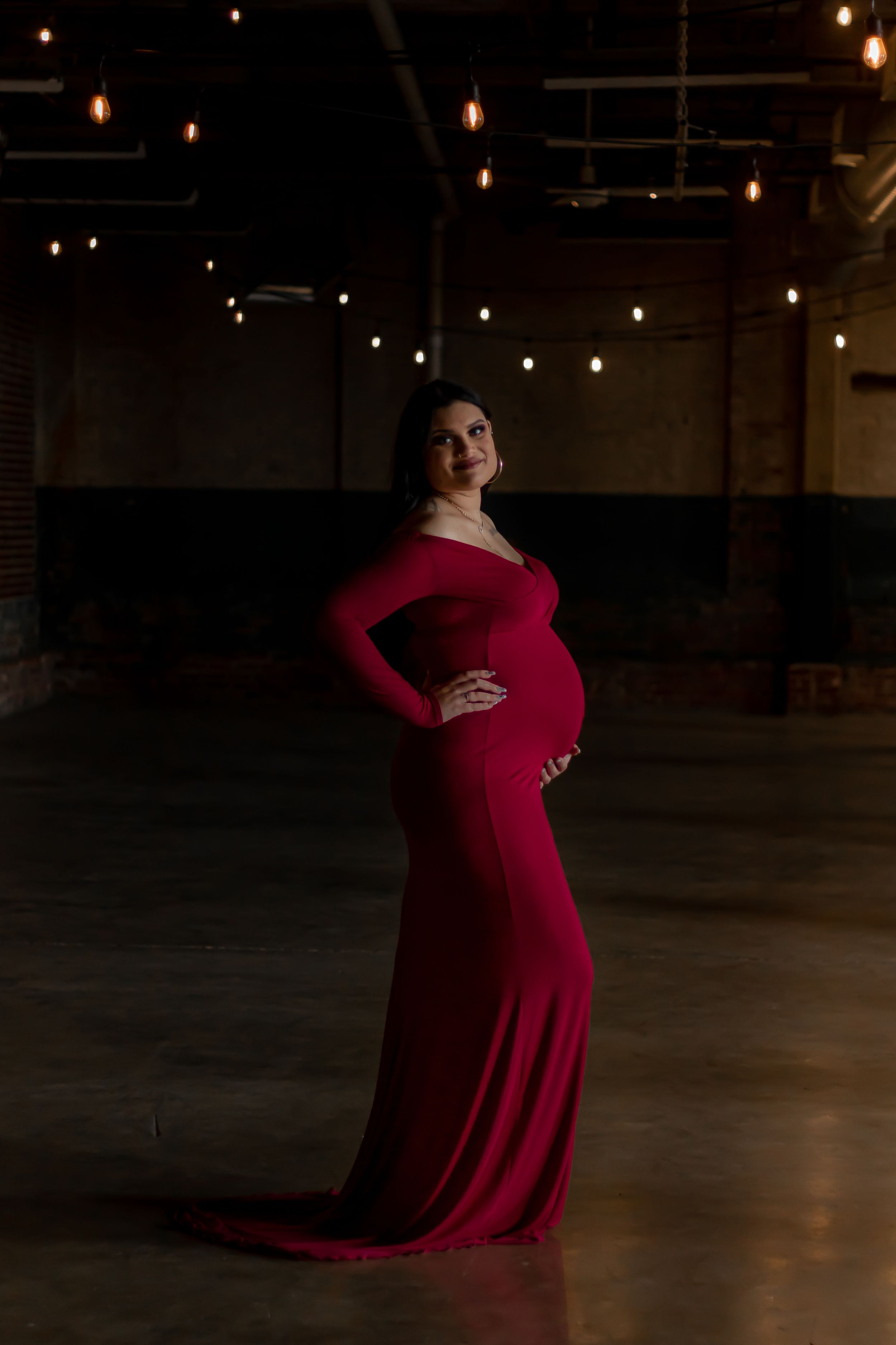 A figure in an elegant red long-sleeve maternity gown poses in a dark atmospheric room with hanging string lights.