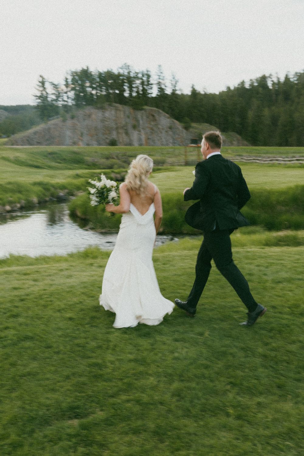 A newlywed couple dances and spins together on grassy terrain near a lake.