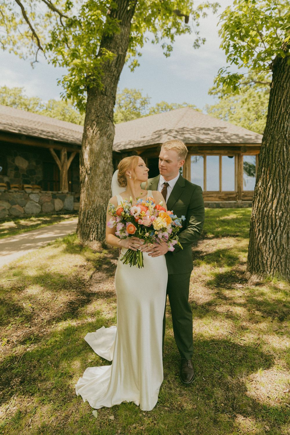 Bride and groom stand together outdoors near trees, she holds a colorful bouquet, wearing a white gown and he a dark suit.
