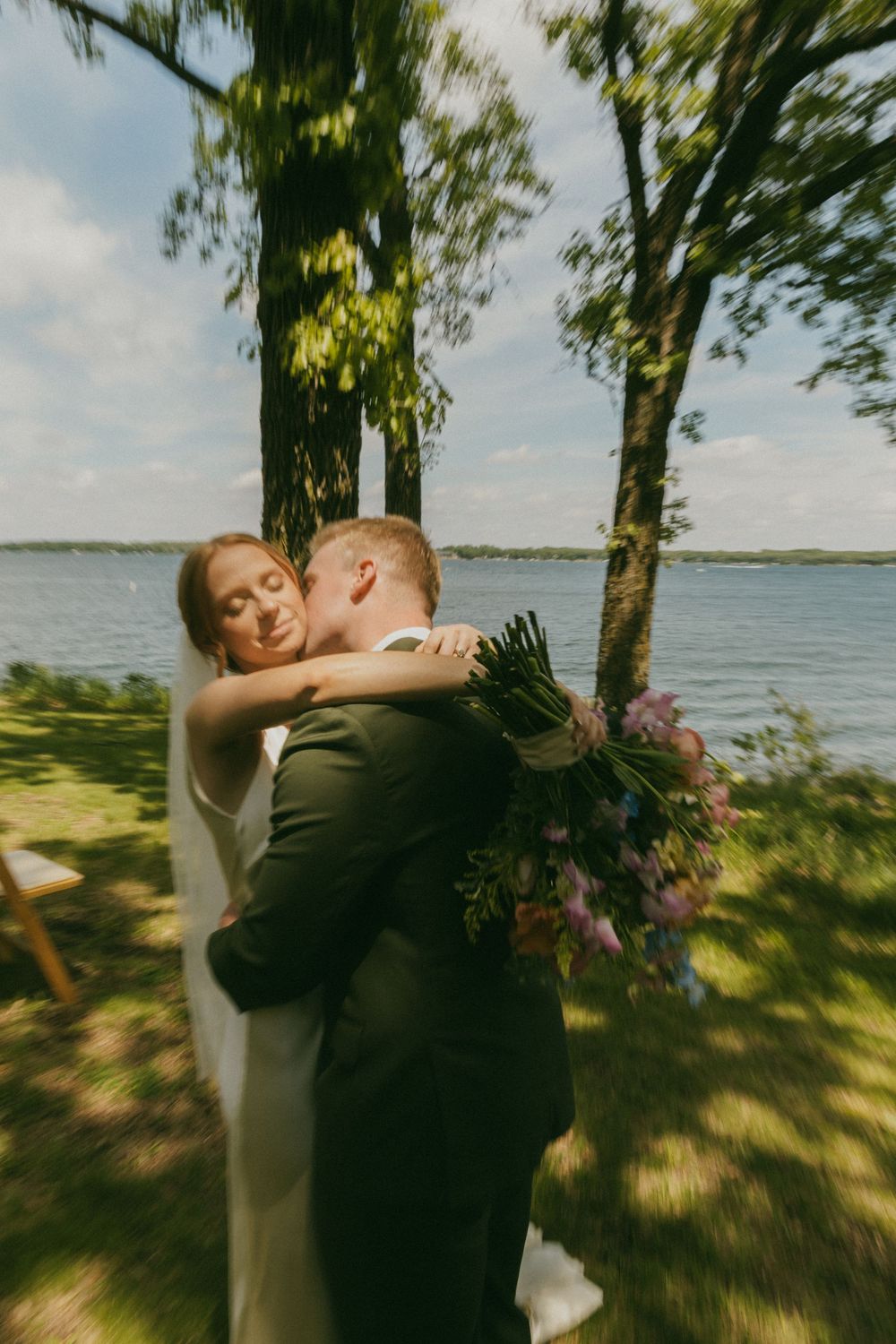 Bride and groom share romantic embrace near lakeside trees, bride holding purple bouquet on sunny wedding day.