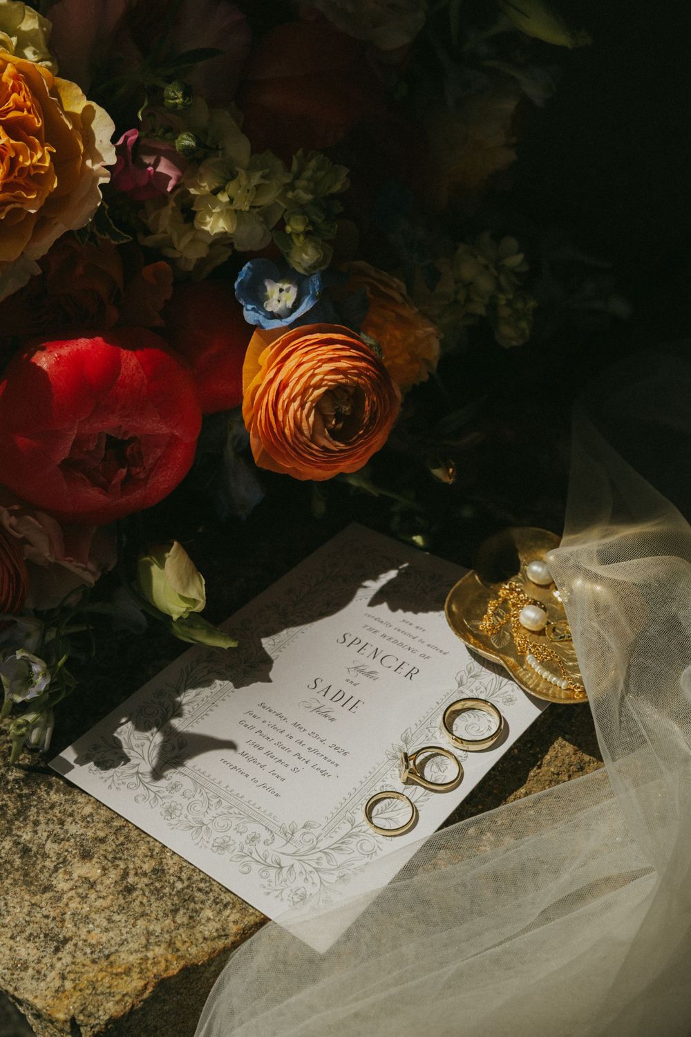 Wedding invitation with gold rings, pearl brooch, and colorful floral bouquet featuring red, orange, and blue blooms.