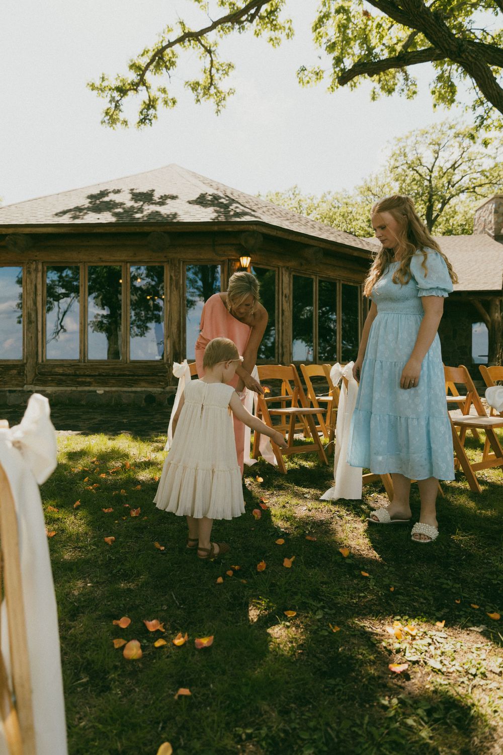 Flower girl scattering petals at outdoor wedding ceremony with wooden chairs and rustic venue backdrop.