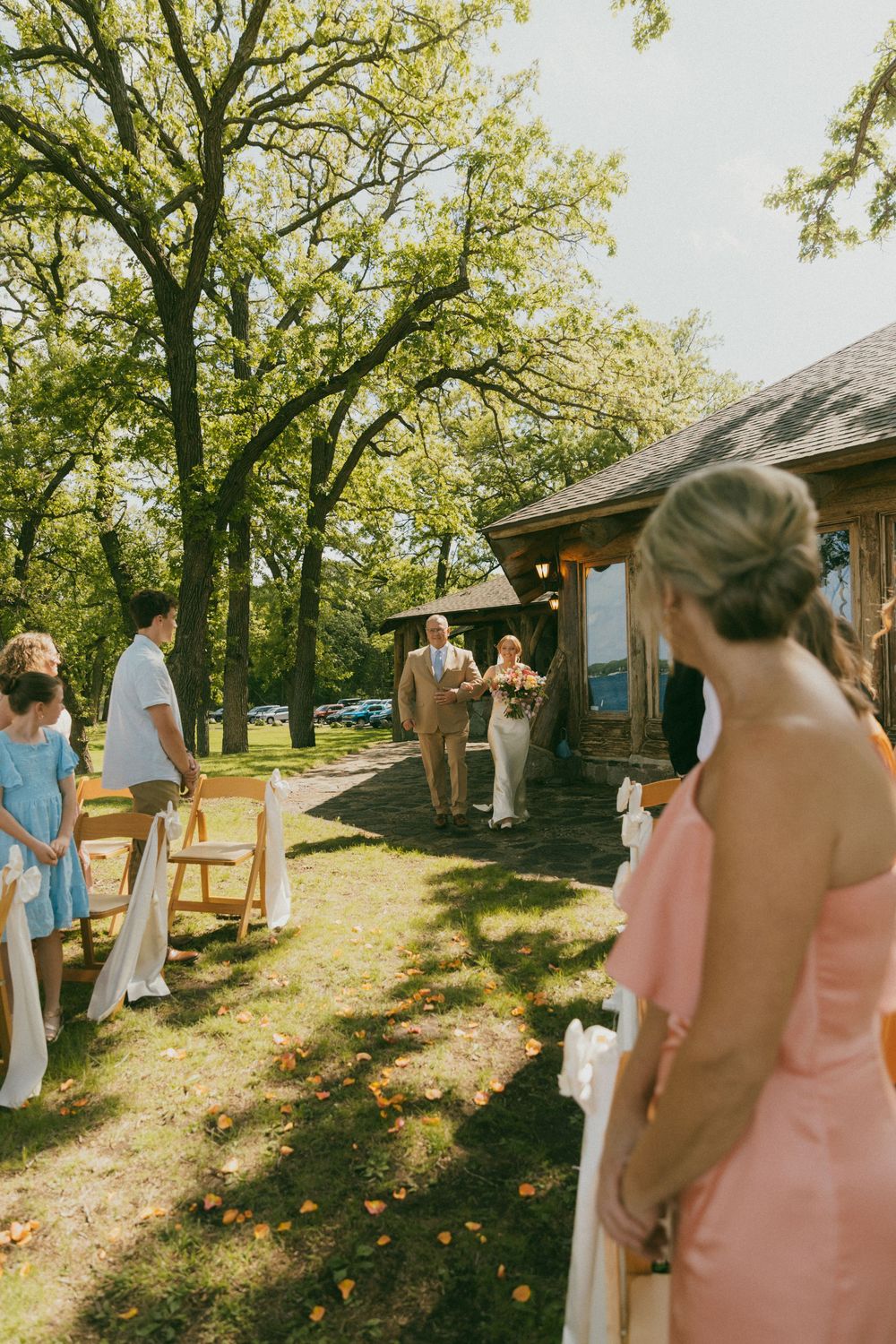 Outdoor wedding ceremony on a sunny day with guests seated on wooden chairs as the bride walks down the aisle.