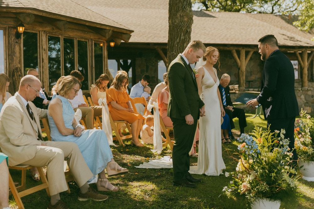 Outdoor wedding ceremony with bride and groom exchanging vows before seated guests near a rustic wooden venue.