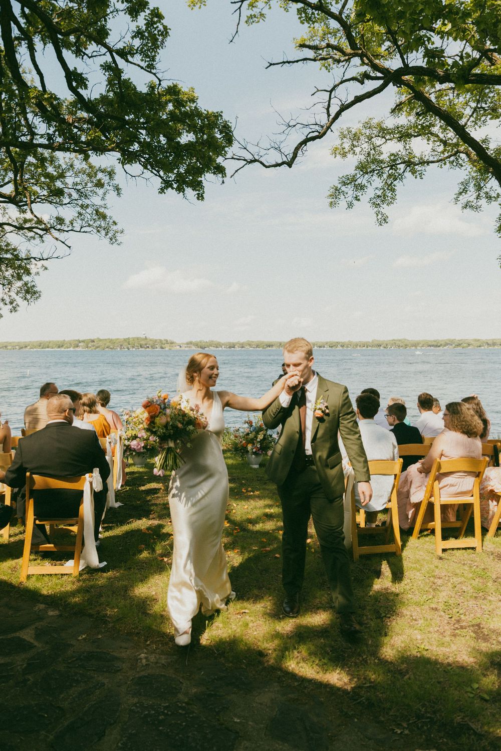 Newlyweds recessional along outdoor lakeside wedding ceremony aisle on sunny day.
