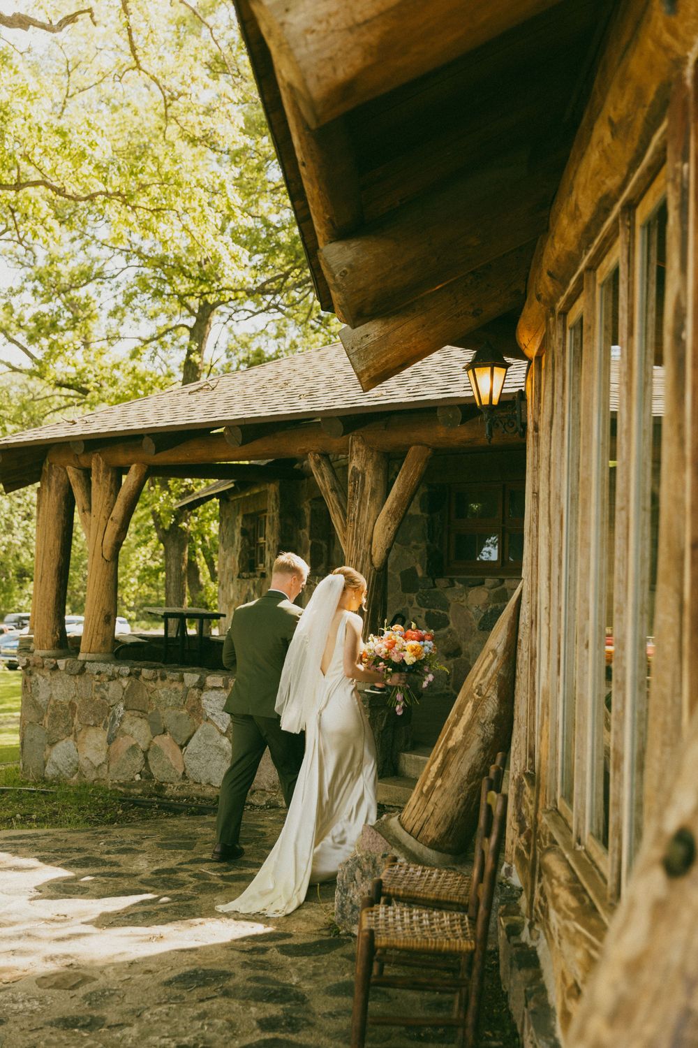 Bride and groom walking along rustic log cabin wedding venue surrounded by lush trees.