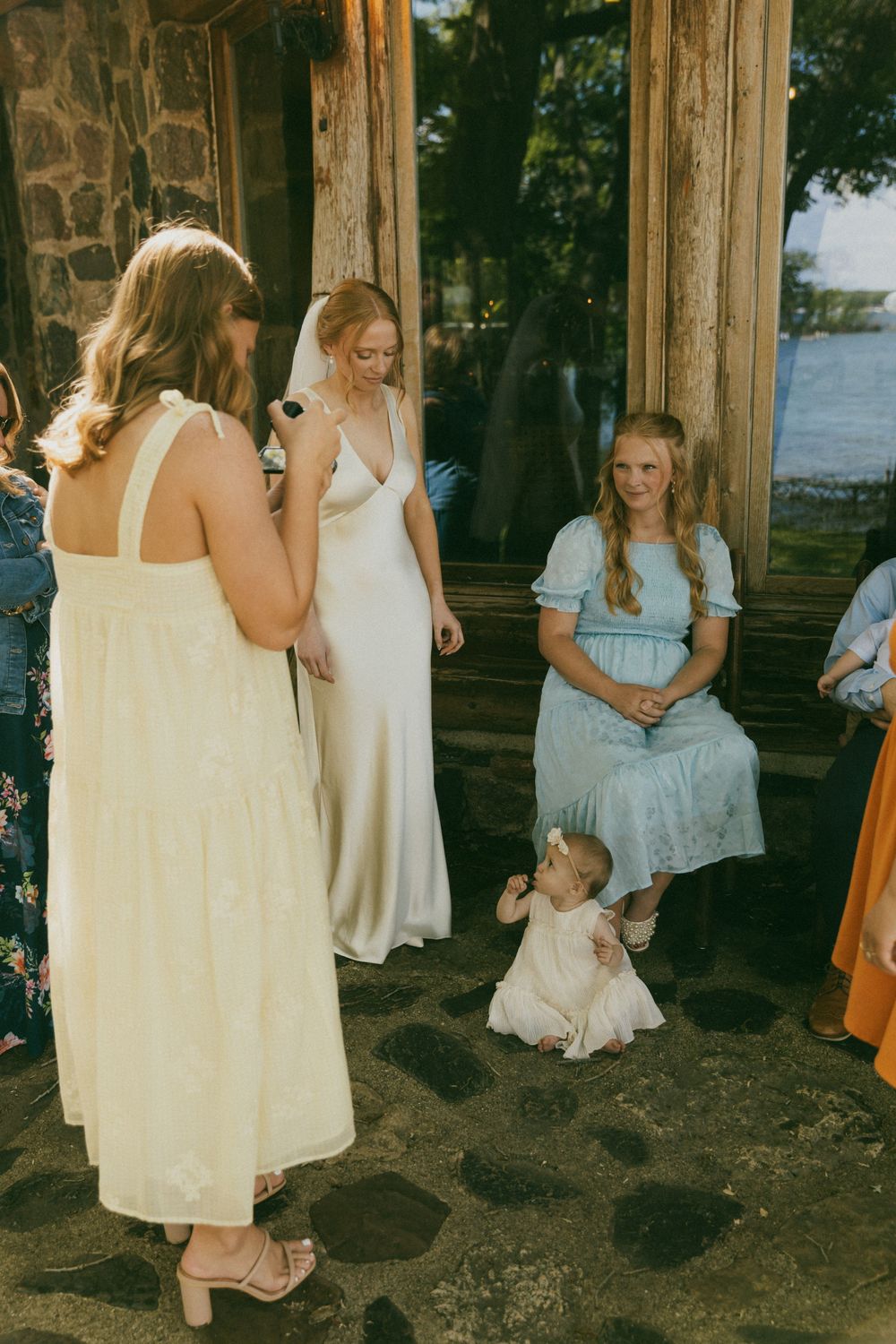 Bride in white satin gown chats with guests in summer dresses near stone building at outdoor wedding reception.