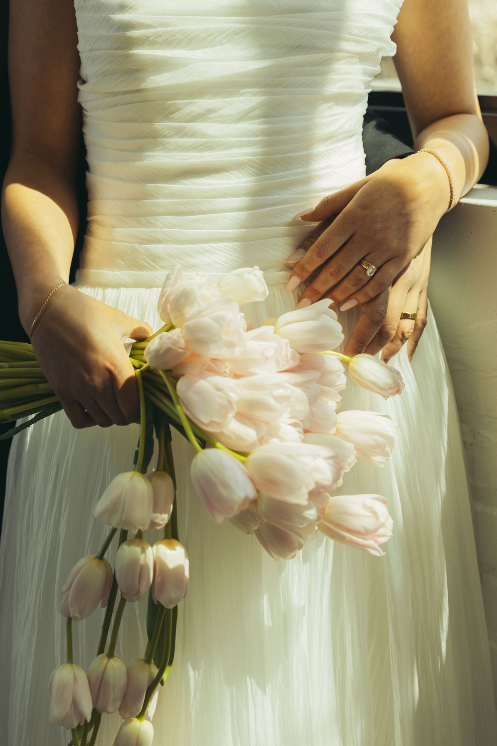 Bride in white dress holding a bouquet of soft pink tulips with wedding rings visible on her hands.