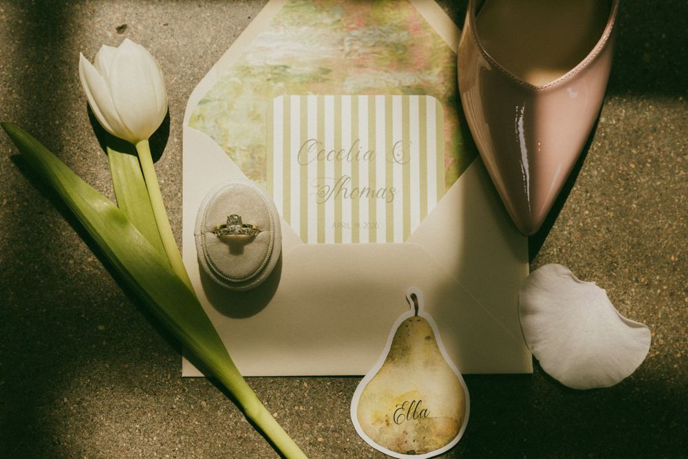 Elegant wedding flat lay with tulip, striped invitation, engagement ring box, pink heels, and pear place card on gold surface.