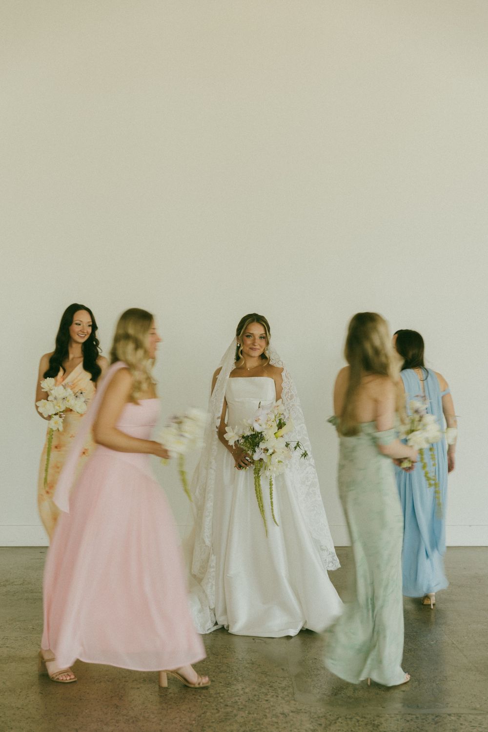 Bride in white gown with veil holds bouquet surrounded by bridesmaids in pastel dresses indoors.