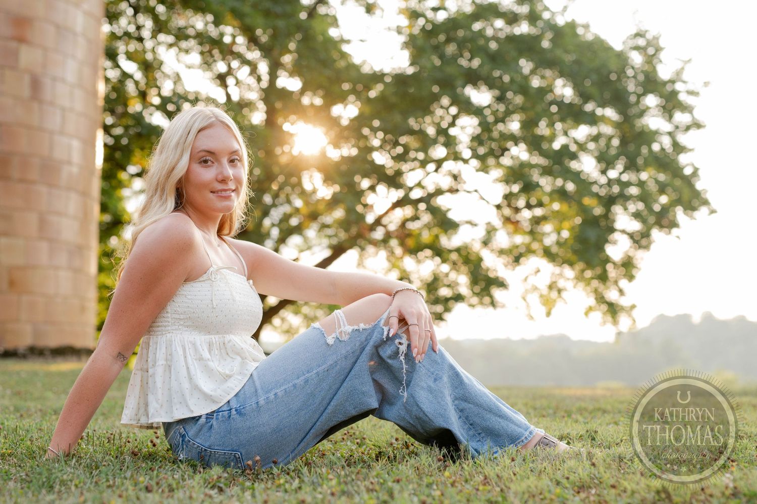 Summer portrait in white top and jeans relaxing on grass during golden hour with trees in background.