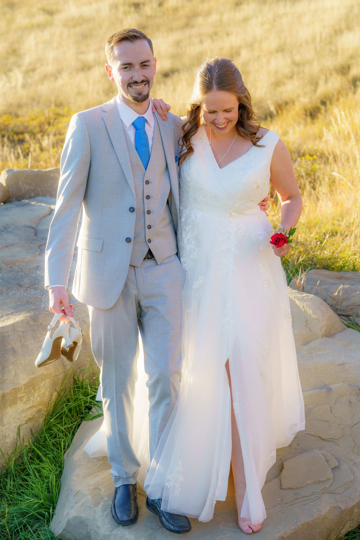 A couple in wedding attire stands on a rocky path with sunlit golden grass in the background at sunset.