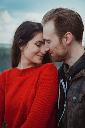 A couple shares an intimate moment wearing red sweater and leather jacket against coastal backdrop.