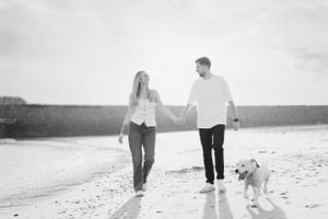 Black and white photo of a couple strolling on the beach with their dog.