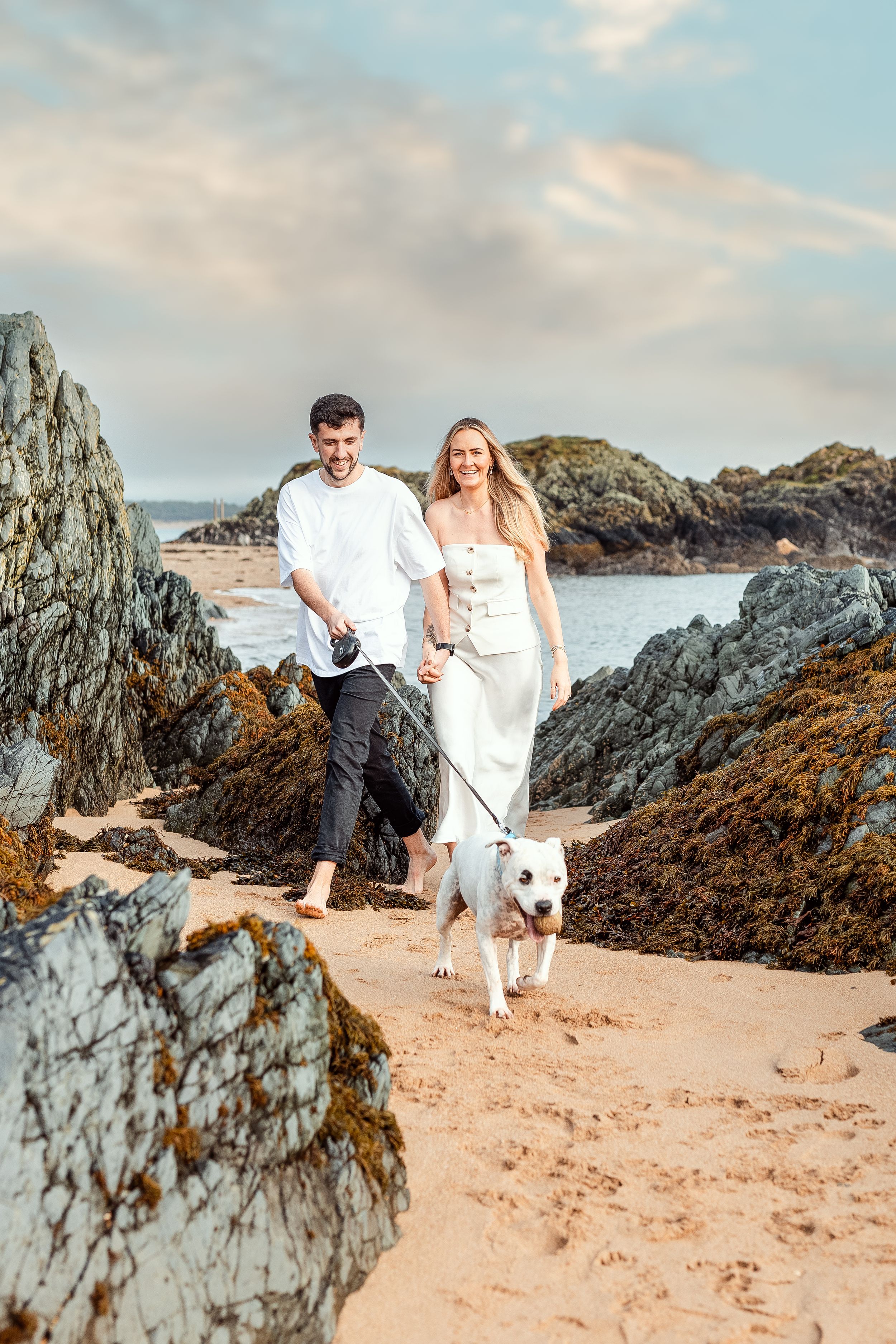 Couple walking their white dog along rocky coastal path with dramatic ocean views.