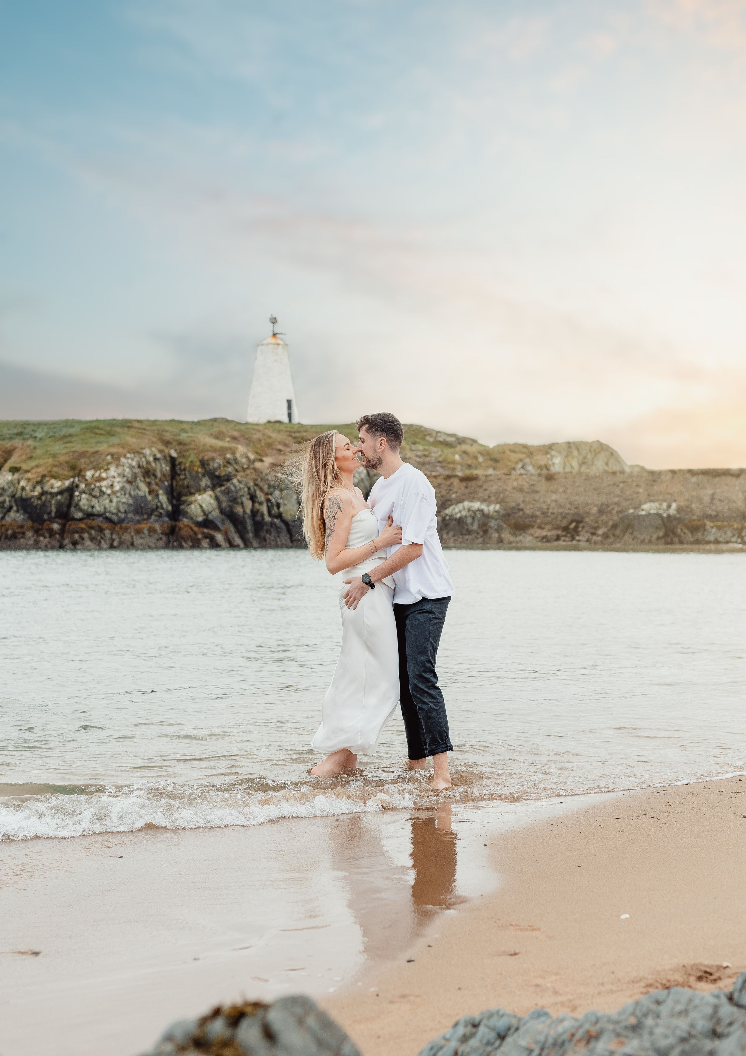 Couple embracing on beach with lighthouse and rocky coastline in background.