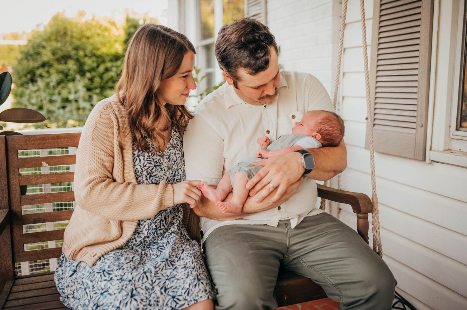 A couple sits together on a porch bench cradling a newborn baby while sharing an intimate family moment.