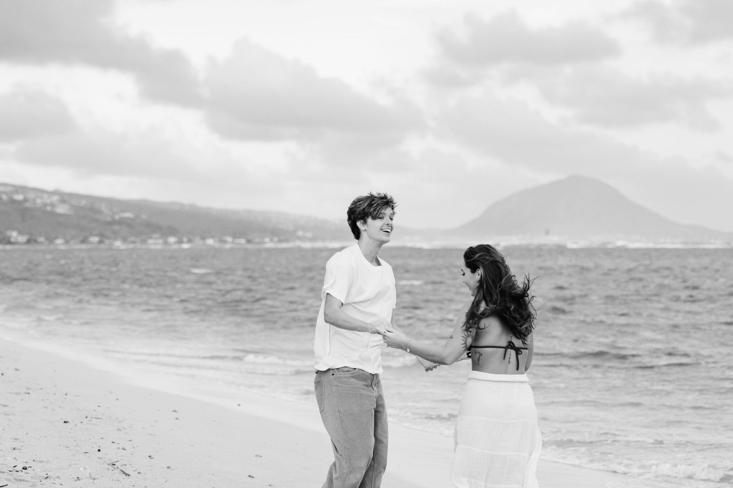 A black and white photo of two people sharing a tender moment on a beach with mountains.