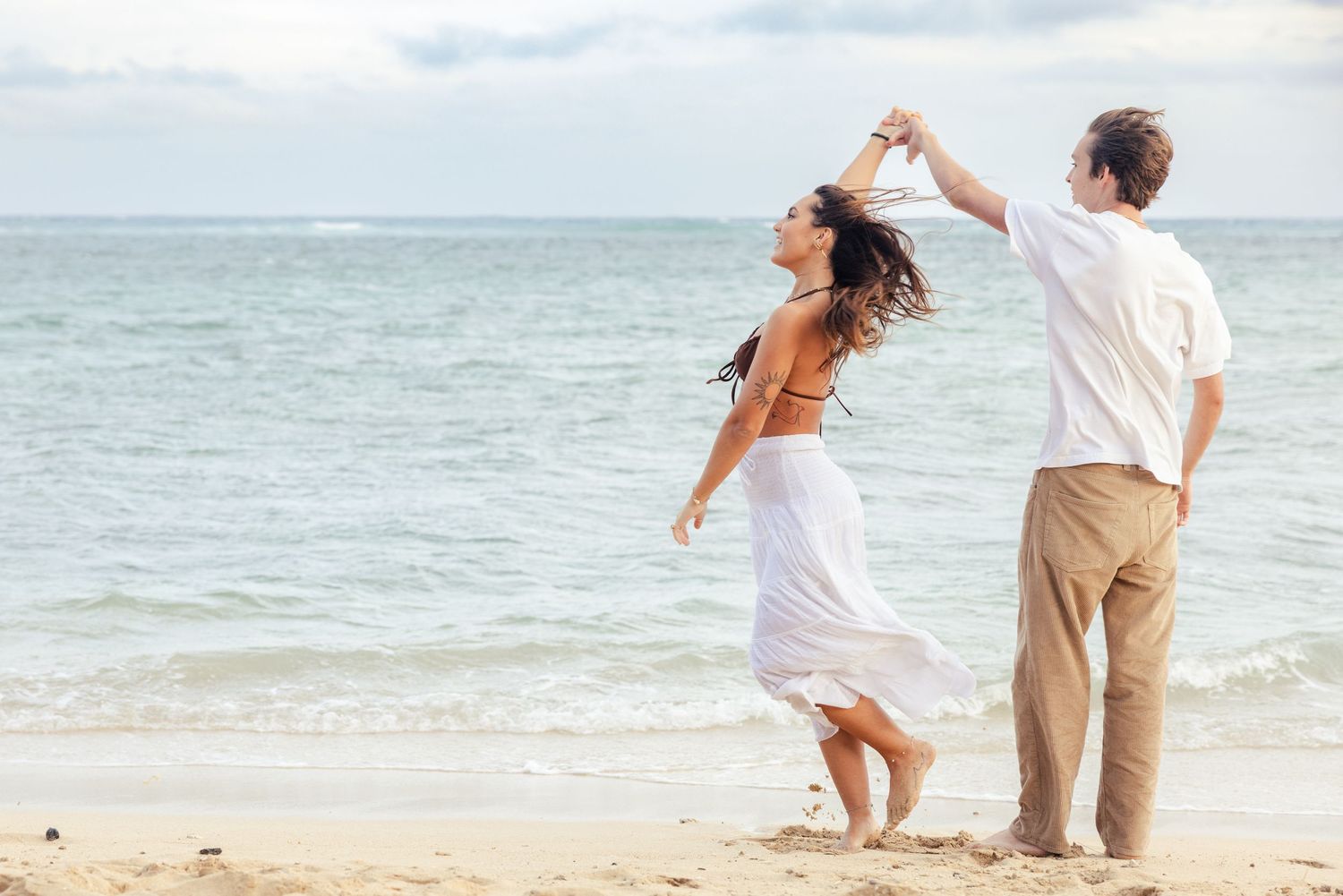 A couple dancing together on a beach with waves and cloudy skies in the background.