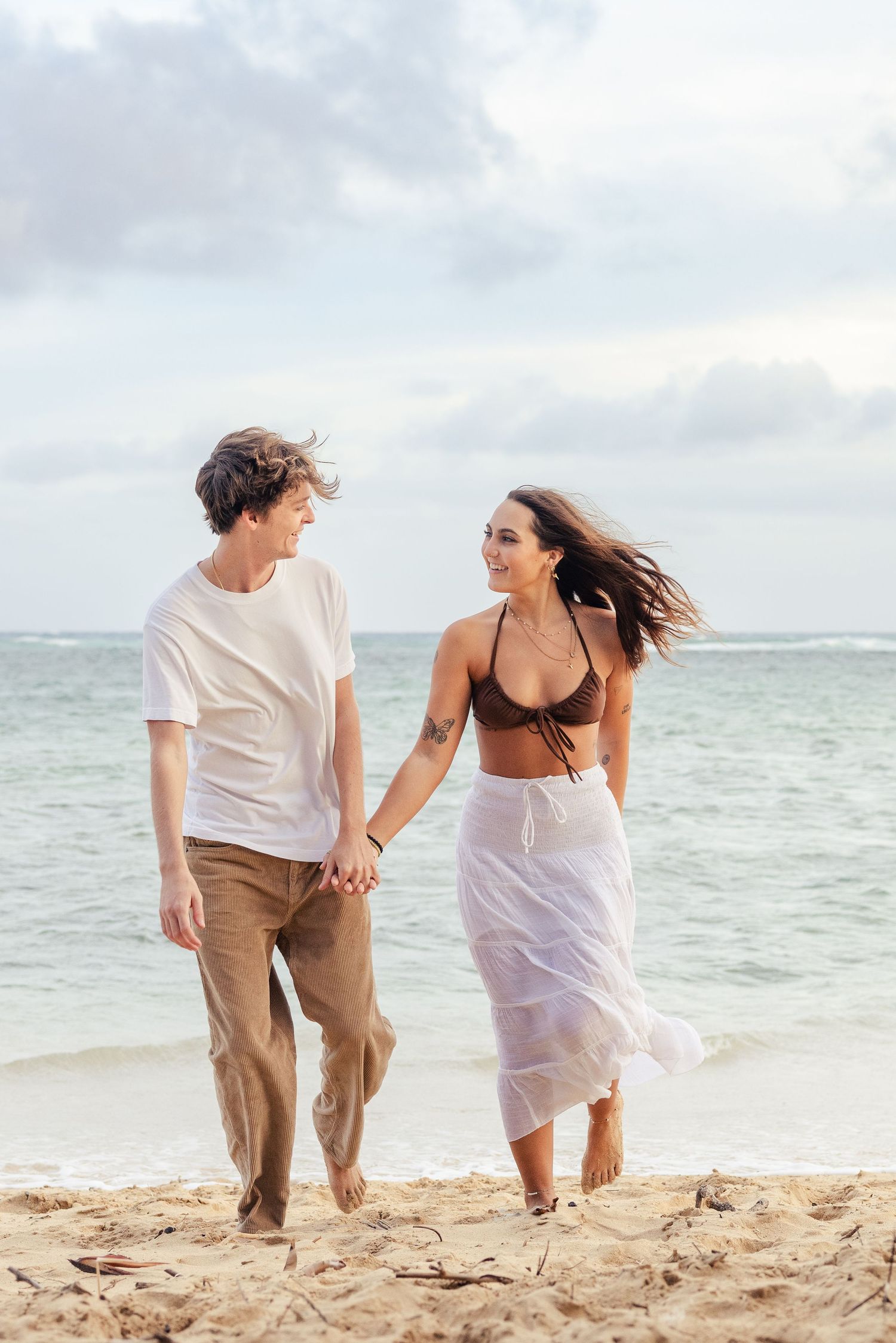A couple walks hand in hand along a tropical beach with turquoise waters and cloudy skies in the background.