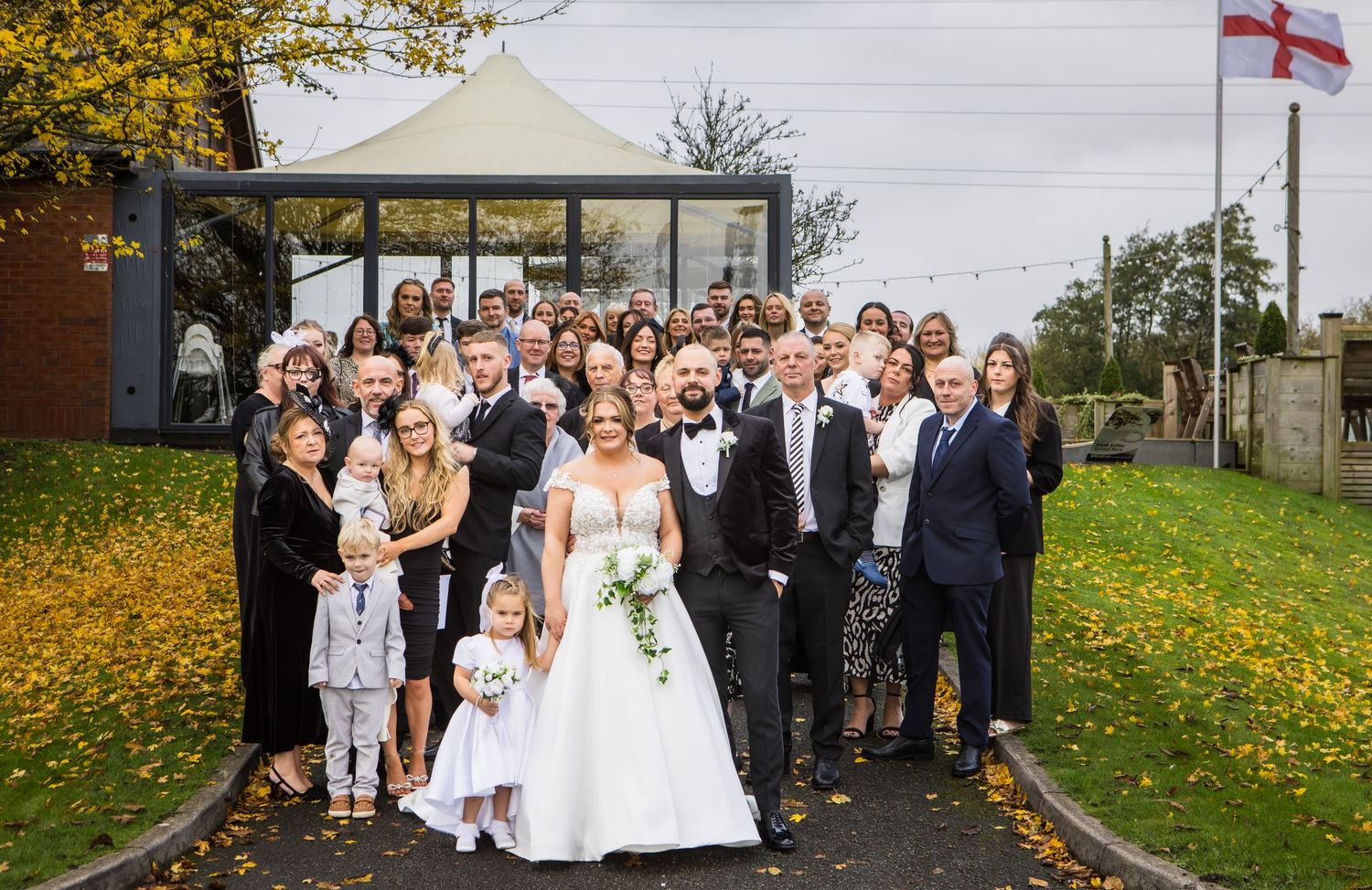Large group wedding photo outdoors with autumn leaves and England flag in background.