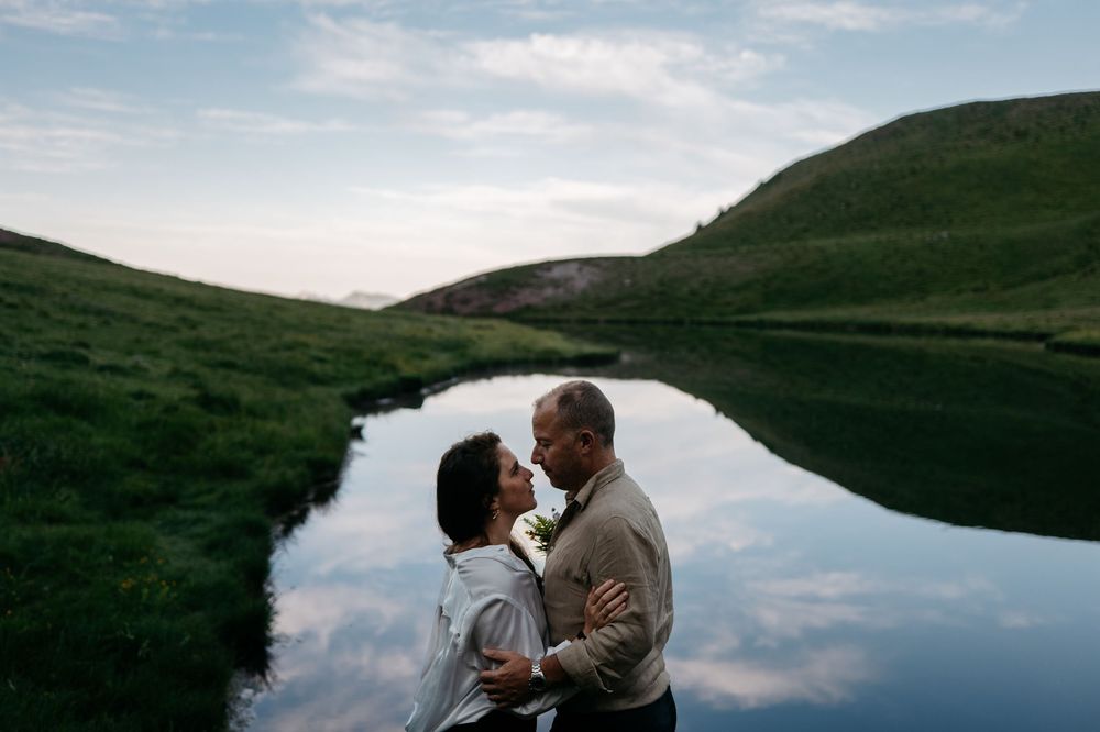 Two silhouetted figures embrace near a tranquil lake with lush green hills and dramatic cloudy skies in the background.