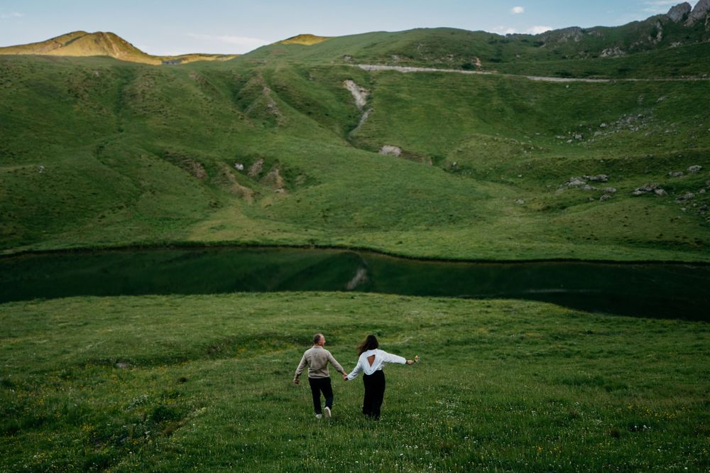 Two figures run hand in hand through a lush green mountain meadow with rolling hills and winding paths in the background.