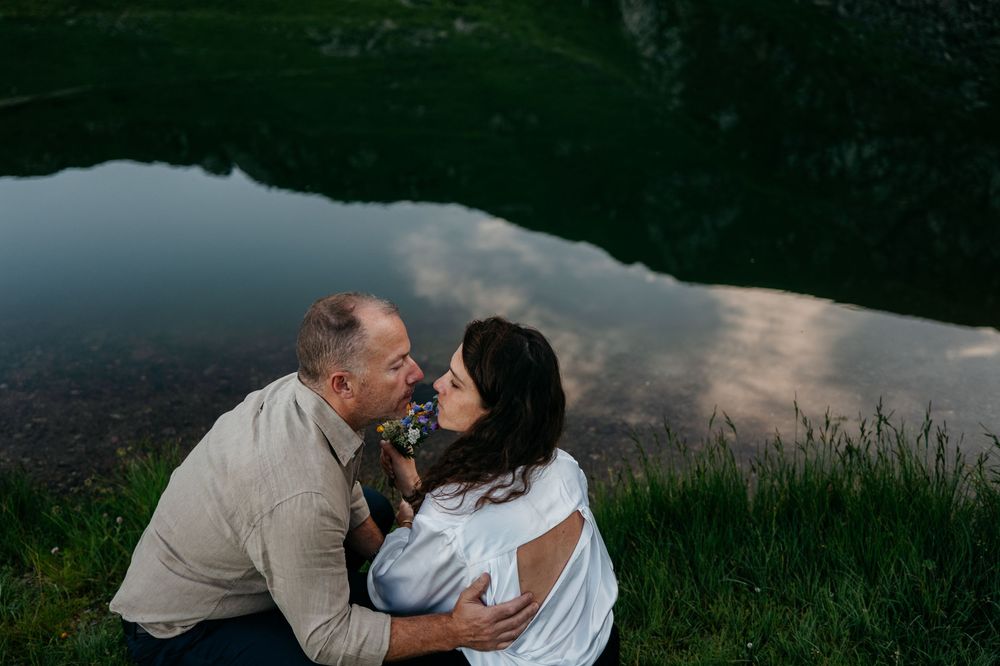 A romantic couple embraces by a serene lake at dusk with reflections of clouds in the calm water.