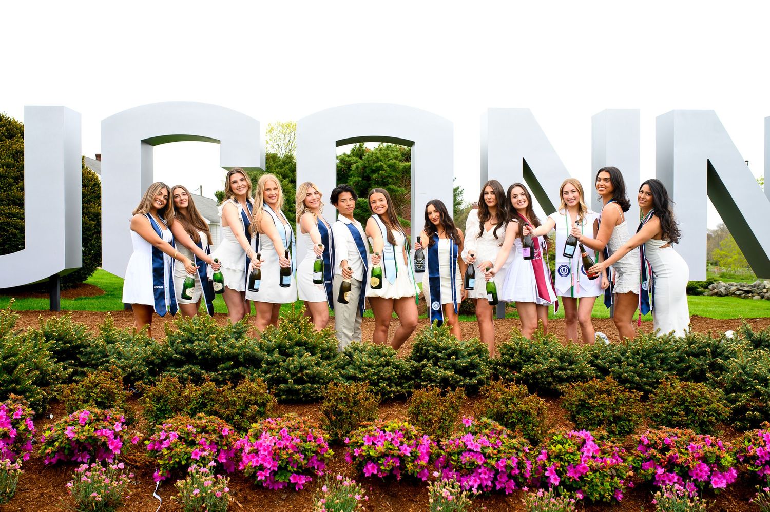 A group in white dresses poses outdoors by large white letters with colorful flowers blooming in the foreground.