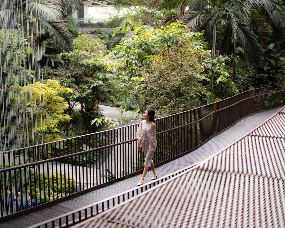 Figure in pink dress walking along elevated walkway surrounded by lush tropical garden.