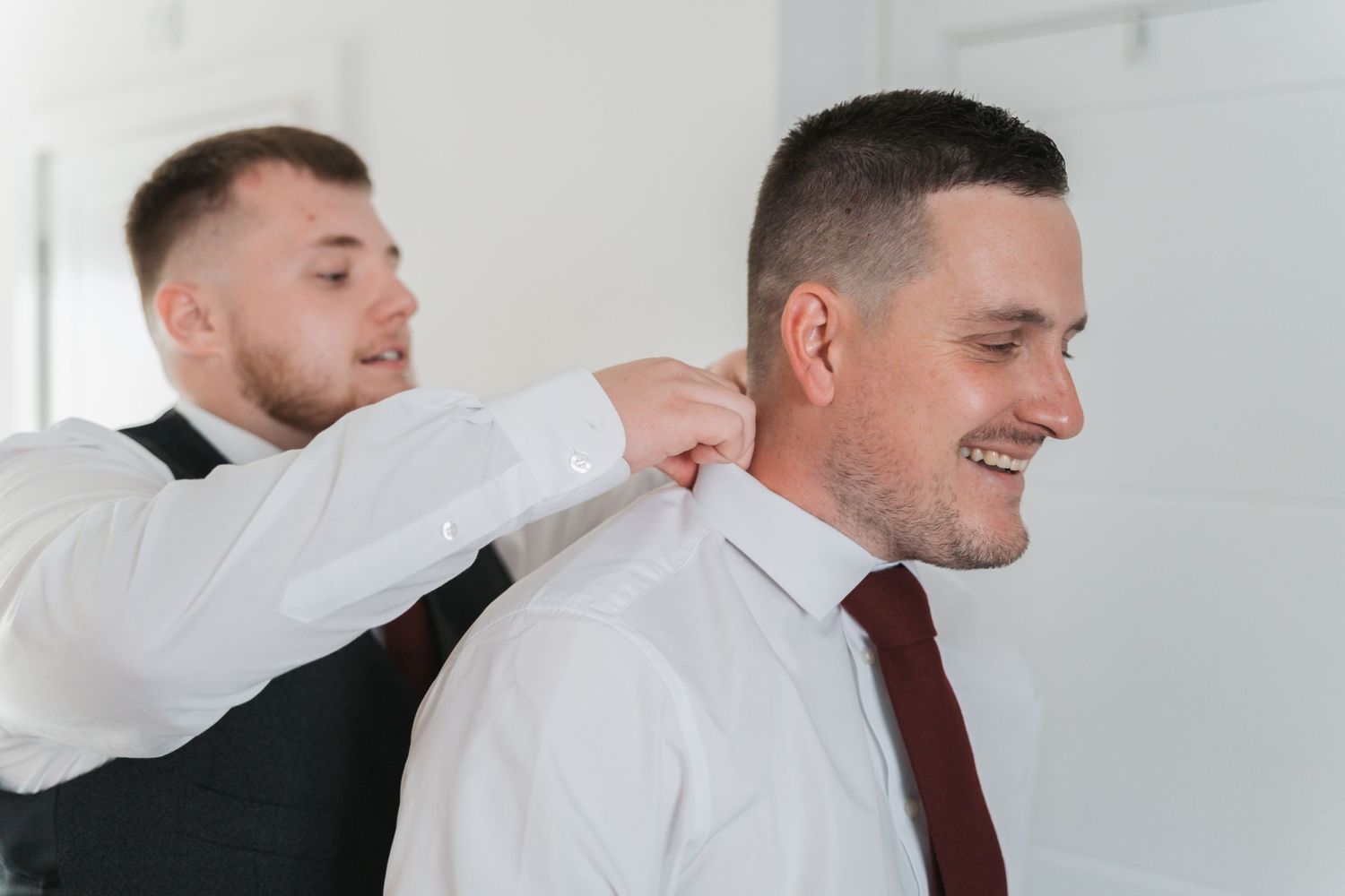 Two people in white dress shirts are indoors during wedding preparations as one helps adjust the other's tie.