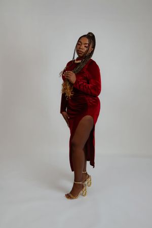 Fashion model in red velvet dress and gold heels poses elegantly against grey background.