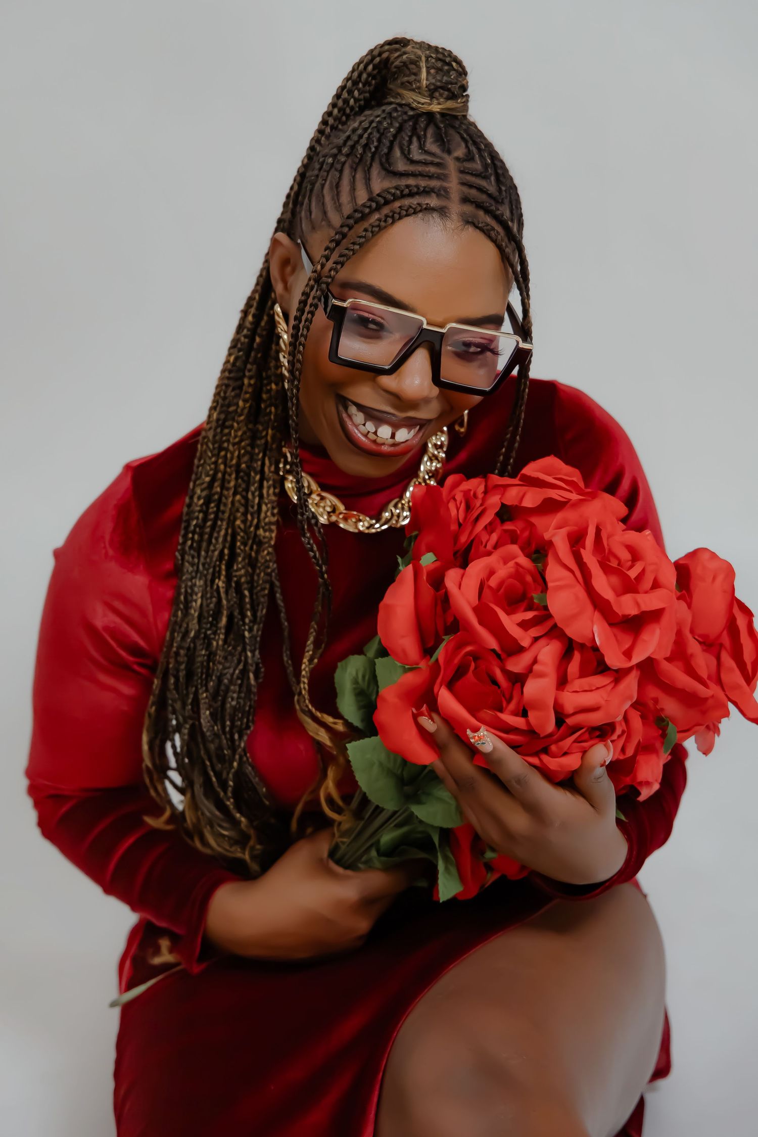 Model with braided hair and glasses holding large red roses while wearing a red dress and gold necklace.