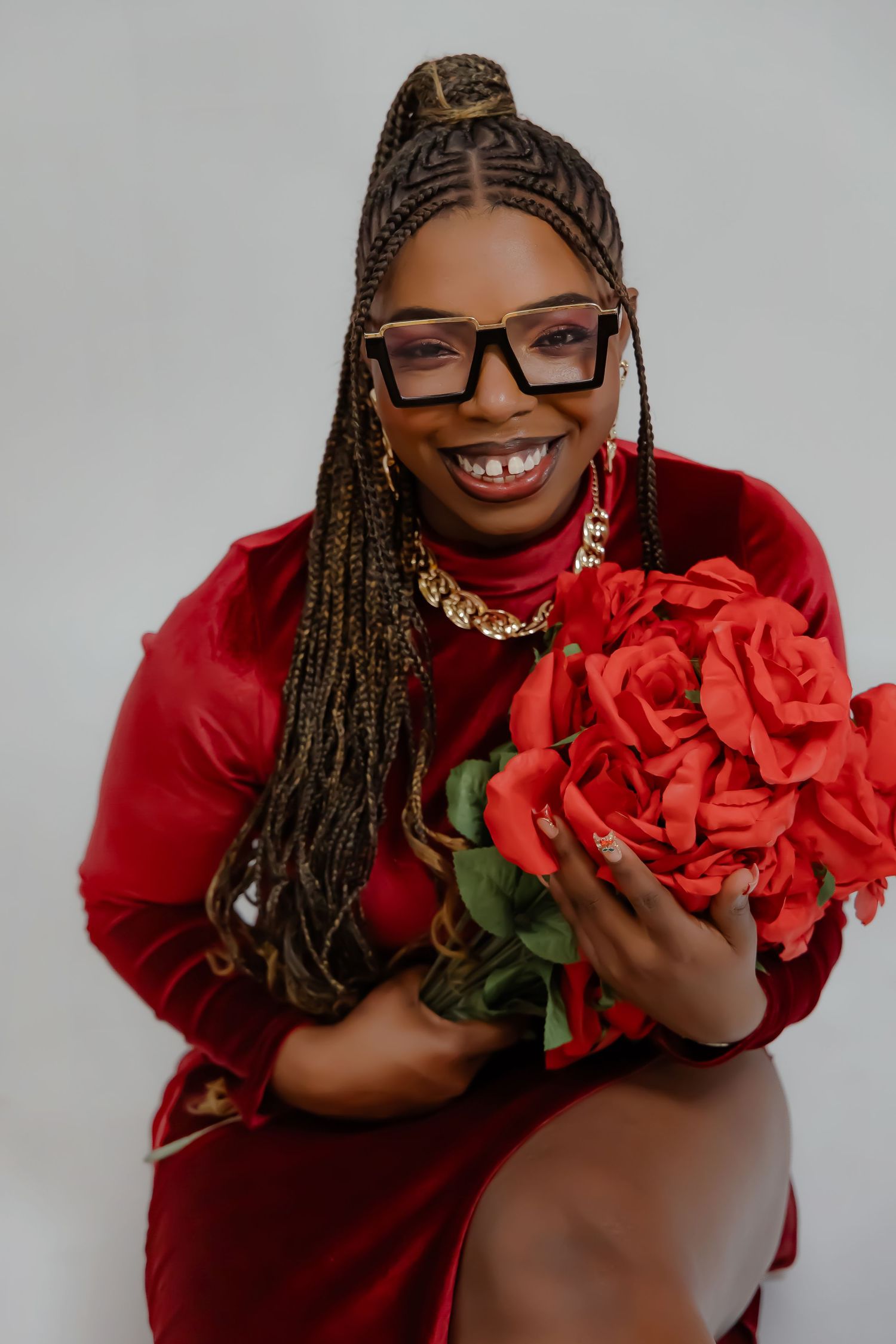 A model in a red velvet dress poses with large red roses while wearing glasses and a gold chain necklace.
