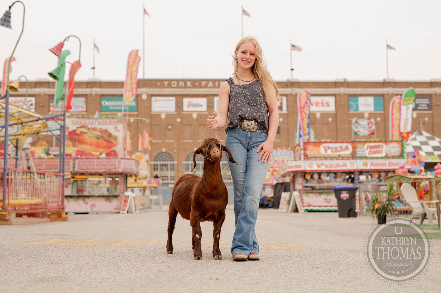 Brown goat stands with handler at county fair with carnival rides in background.