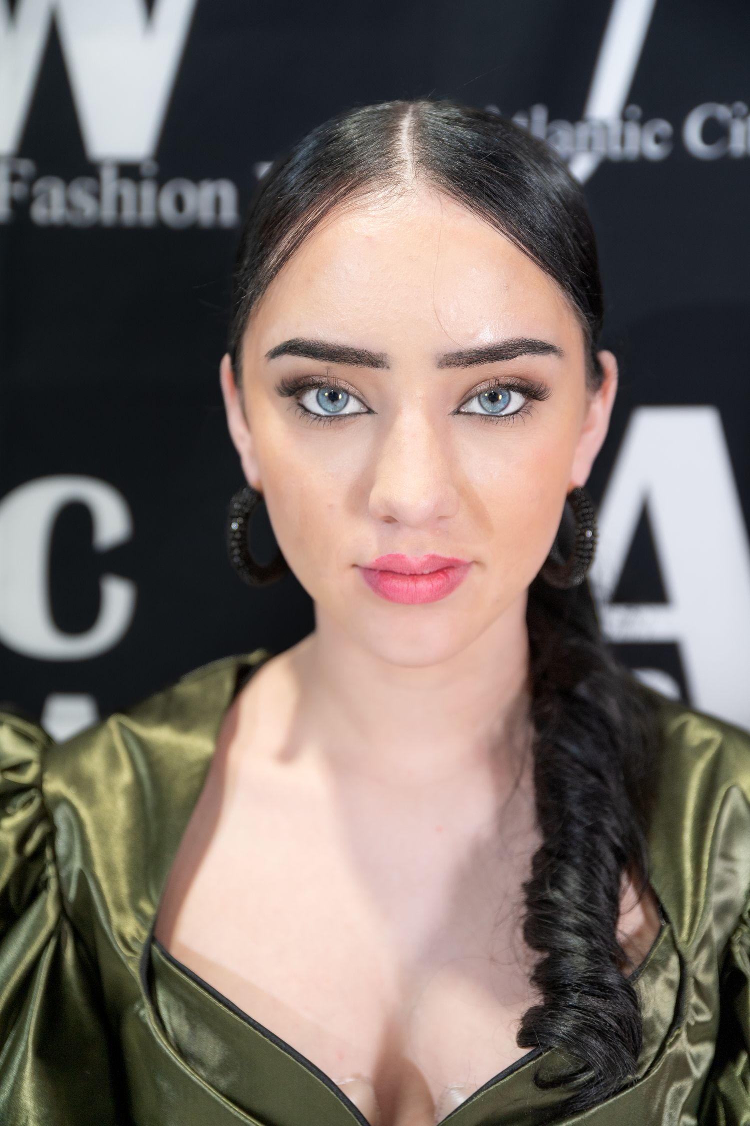 Portrait of a woman with striking eyes and dark braided hair wearing an olive green metallic jacket against a black background.