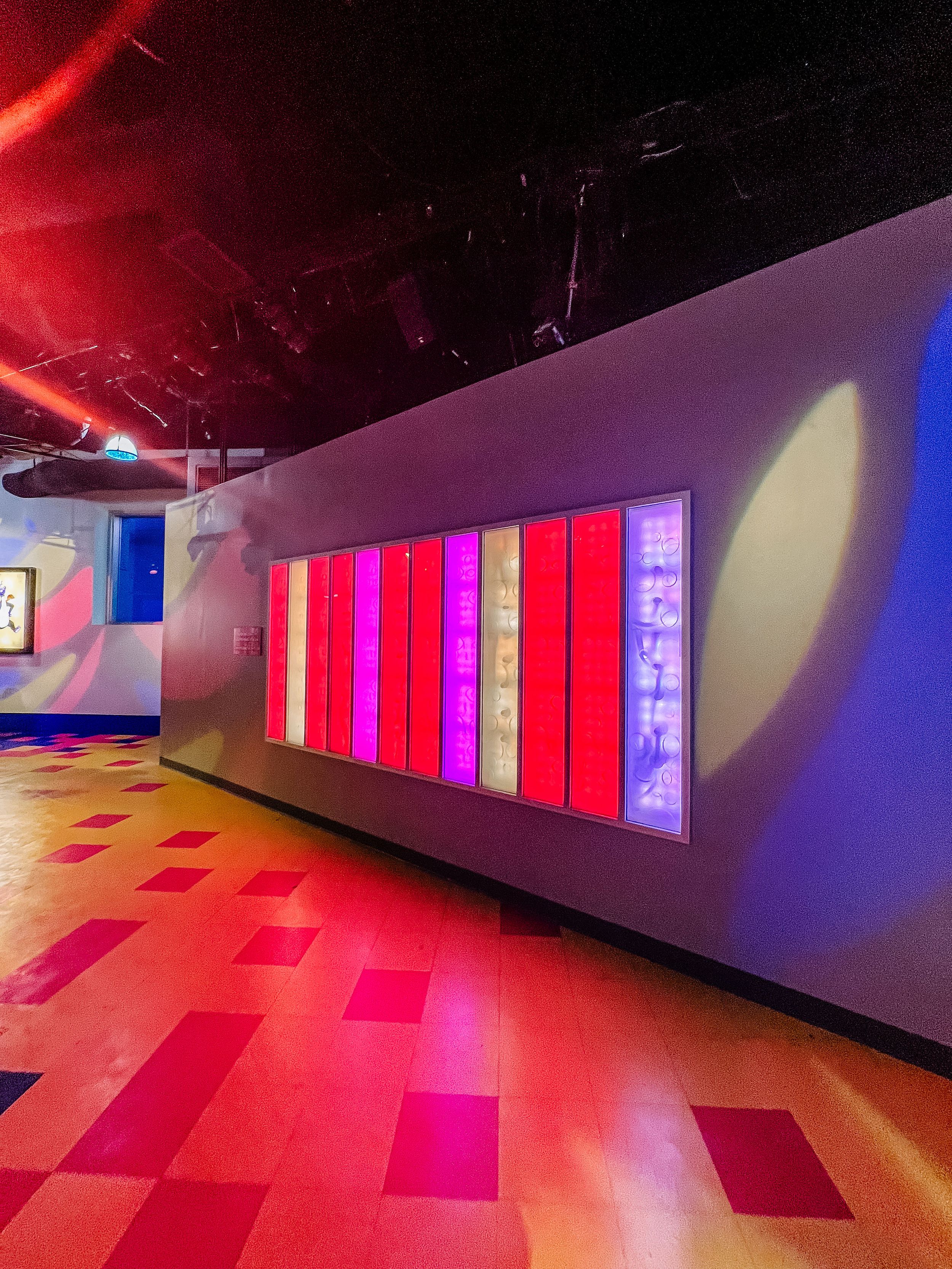Modern hallway with colorful LED wall panels and geometric patterned floor illuminated in vibrant pink and red lights.